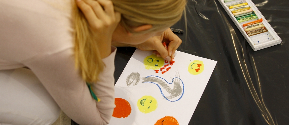 Zdjęcie: blondwłosa dziewczynka pochylona nad rysunkiem, który tworzy pastelami. 