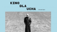 Czaro-białe zdjęcie Chinasky'ego. Stoi na trawniku w długim, sięgającym ziemi czarnym płaszczu i czarnych okularach. Ręce w kieszeniach płaszcza