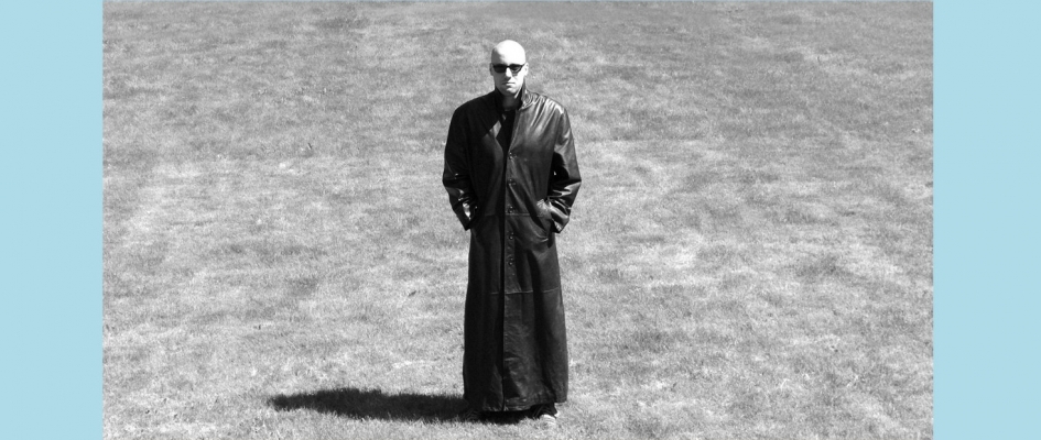 Czaro-białe zdjęcie Chinasky'ego. Stoi na trawniku w długim, sięgającym ziemi czarnym płaszczu i czarnych okularach. Ręce w kieszeniach płaszcza