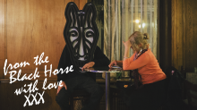 Dwie osoby siedzą przy niewielkim kawiarnianym stoliku. Kobieta pochylona nad telefonem komórkowym, druga osoba ma na głowie ogromną maskę konia